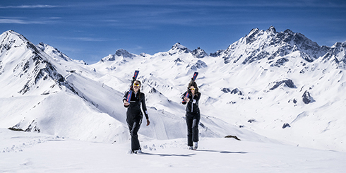 Due persone che portano gli sci camminano sulla neve con le montagne sullo sfondo sotto un cielo azzurro.